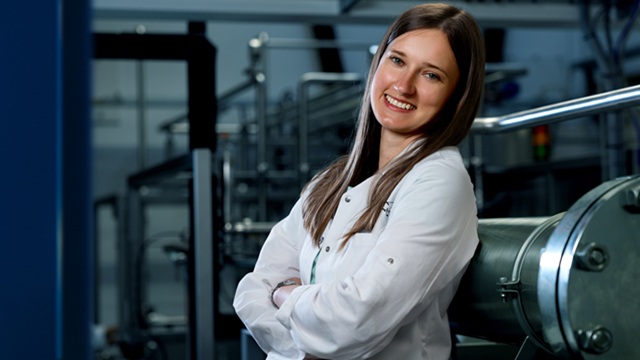Person in a lab coat stands with arms crossed in an industrial or laboratory setting, face blurred
