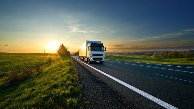 A white semi‑truck driving along a rural highway at sunset, surrounded by open fields and warm golden light, representing long‑haul transportation and logistics
